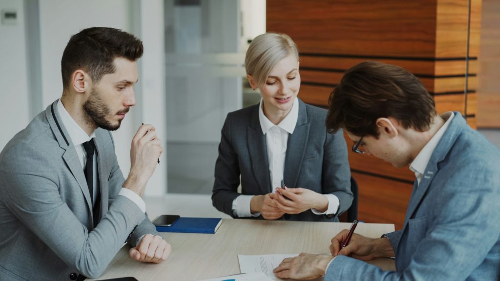 Three professionals in gray suits sit at a table reviewing documents in a modern office setting.