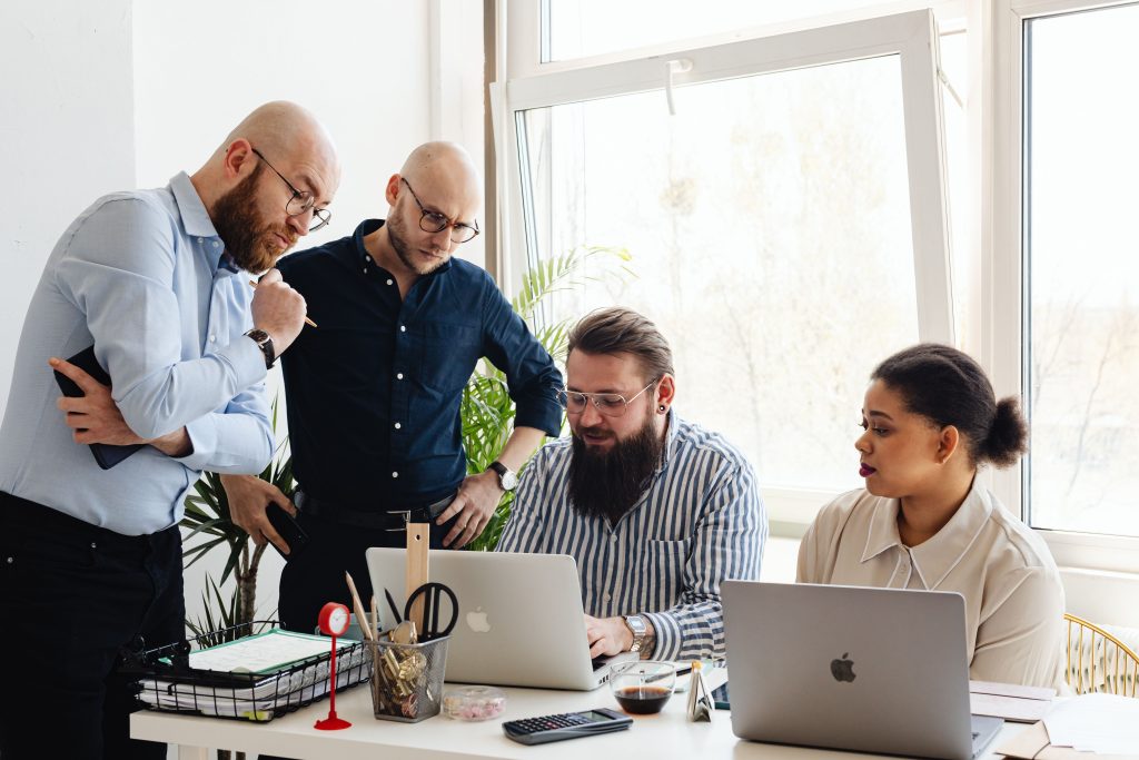 Five coworkers collaborate around a desk with laptops in a bright office by a window, discussing a project).