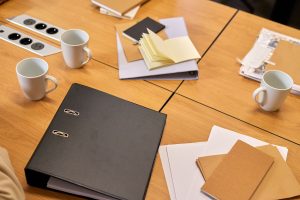 A black binder sits on a wooden table with scattered papers, envelopes, and a few white mugs nearby, suggesting a workspace.