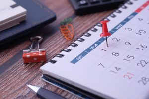 Close-up of a desk with a calendar page and a red pushpin marking a date, with stationery in the background.