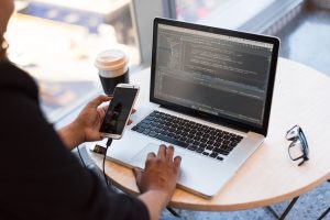 Person typing on a laptop with code visible on screen, while holding a connected smartphone at a round table with coffee and glasses nearby
