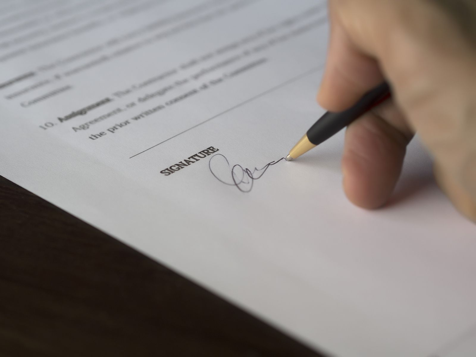 Close-up of a hand signing a document, pen writing a signature on the SIGNATURE line on a contract surface.