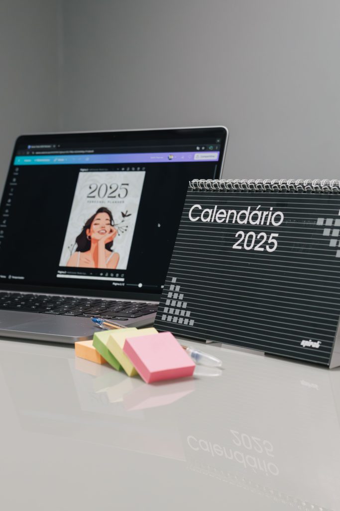 Laptop on a desk showing a calendar design and a spiral-bound notebook labeled 'Calendário 2025' beside colorful sticky notes.