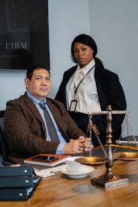 Two professionals in a law office: a man seated at a desk with papers, a phone, and a scale of justice nearby, while a woman in a blazer stands beside him wearing an ID badge.