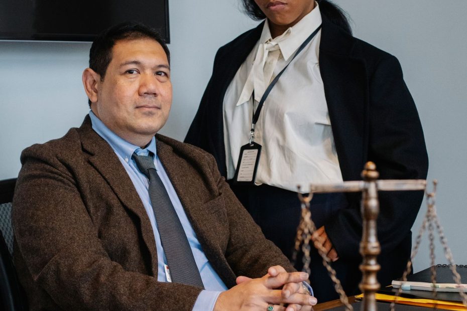 Two professionals in a law office: a man seated at a desk with papers, a phone, and a scale of justice nearby, while a woman in a blazer stands beside him wearing an ID badge.