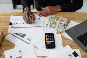 Hands writing on financial documents with cash, a calculator, and a laptop on a wooden desk