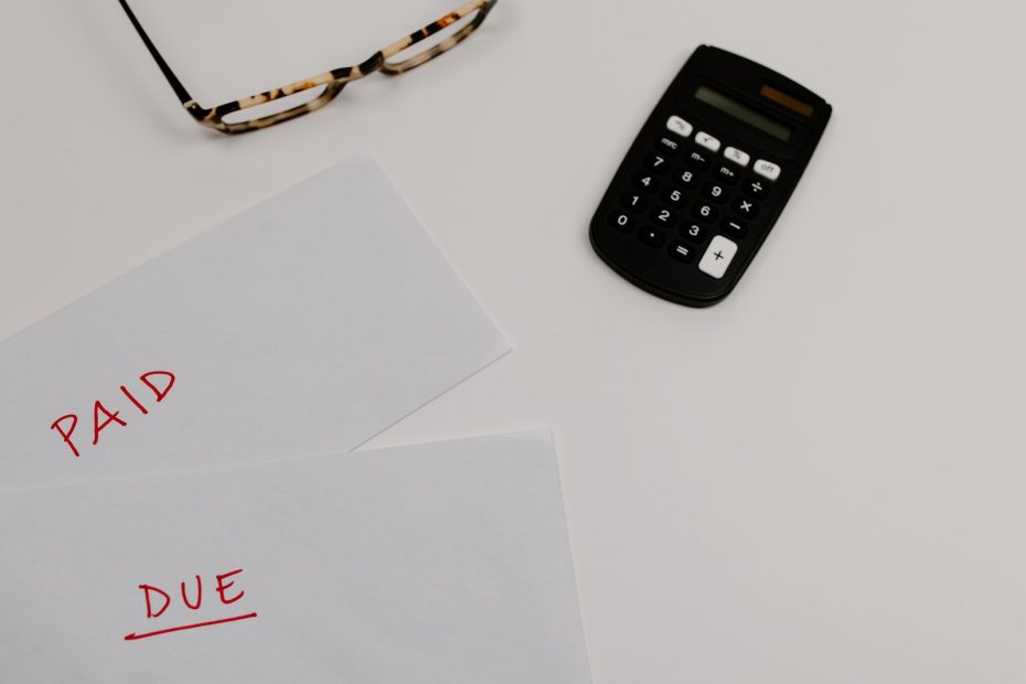 Desk scene with two white envelopes showing red handwritten 'PAID' and 'DUE' labels, plus a pair of tortoiseshell glasses and a black calculator on a white surface.