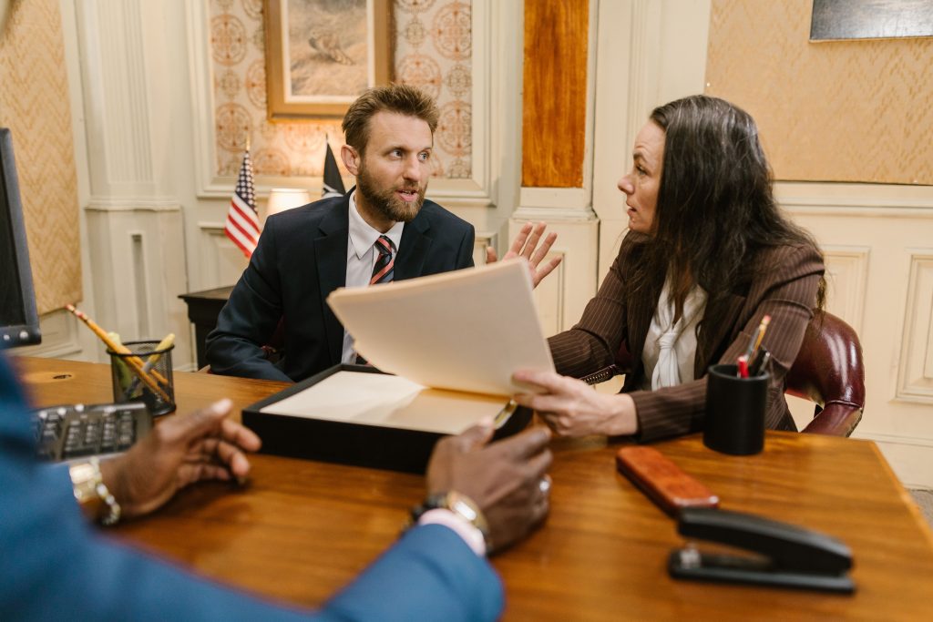 Two professionals discuss documents at a wooden table; man in a suit talks to a woman holding papers in a formal office setting with flags in the background.