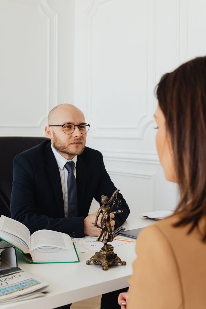 Man in a suit and glasses sits at a desk across from a client, with open books and a small statue on the desk in a bright office.