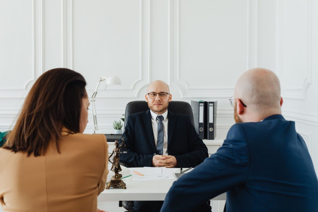 Three professionals sit around a conference desk in a bright office, discussing documents and taking notes.