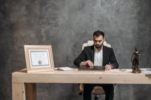 Man in a suit sits at a wooden desk, working on a laptop with a framed certificate, open notebook, and a Lady Justice statue nearby against a gray wall.