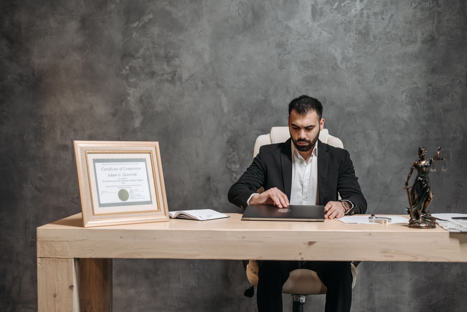 Professional man in a dark suit sits at a wooden desk, working on a laptop with a framed certificate and a statue of justice nearby on the desk