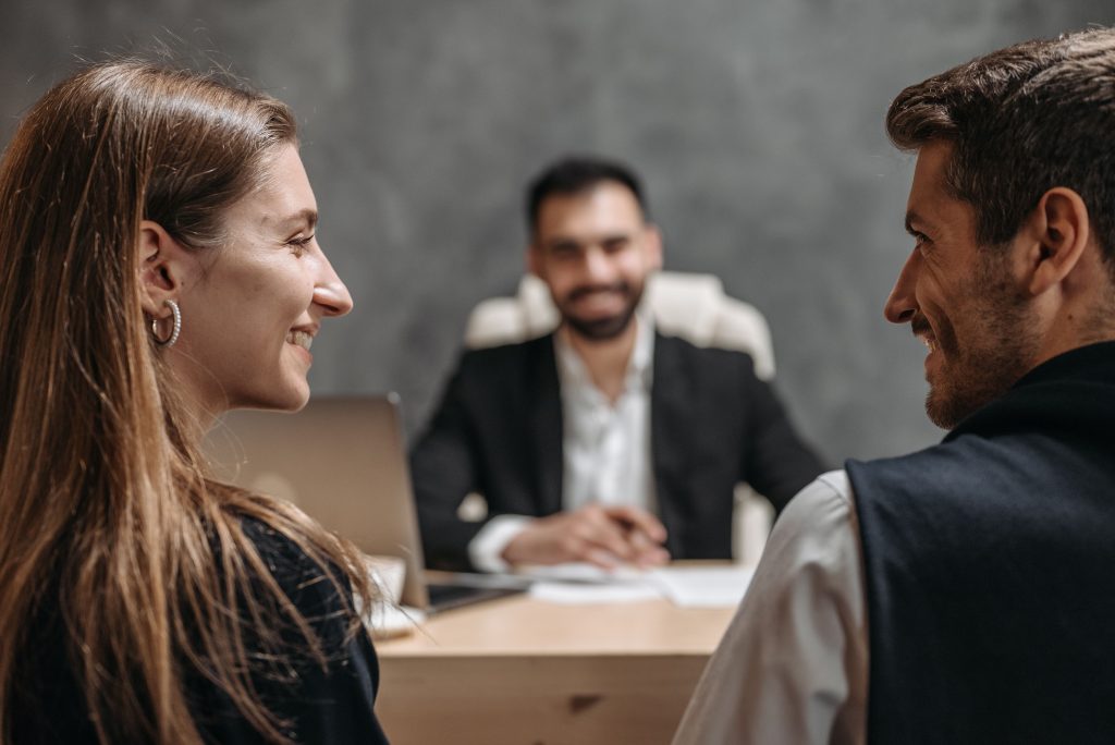 Two coworkers smile and talk in a meeting, with a blurred colleague seated at a desk in the background.