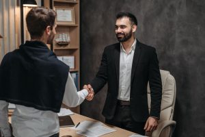Two men in business attire shake hands across a desk in a modern office setting.