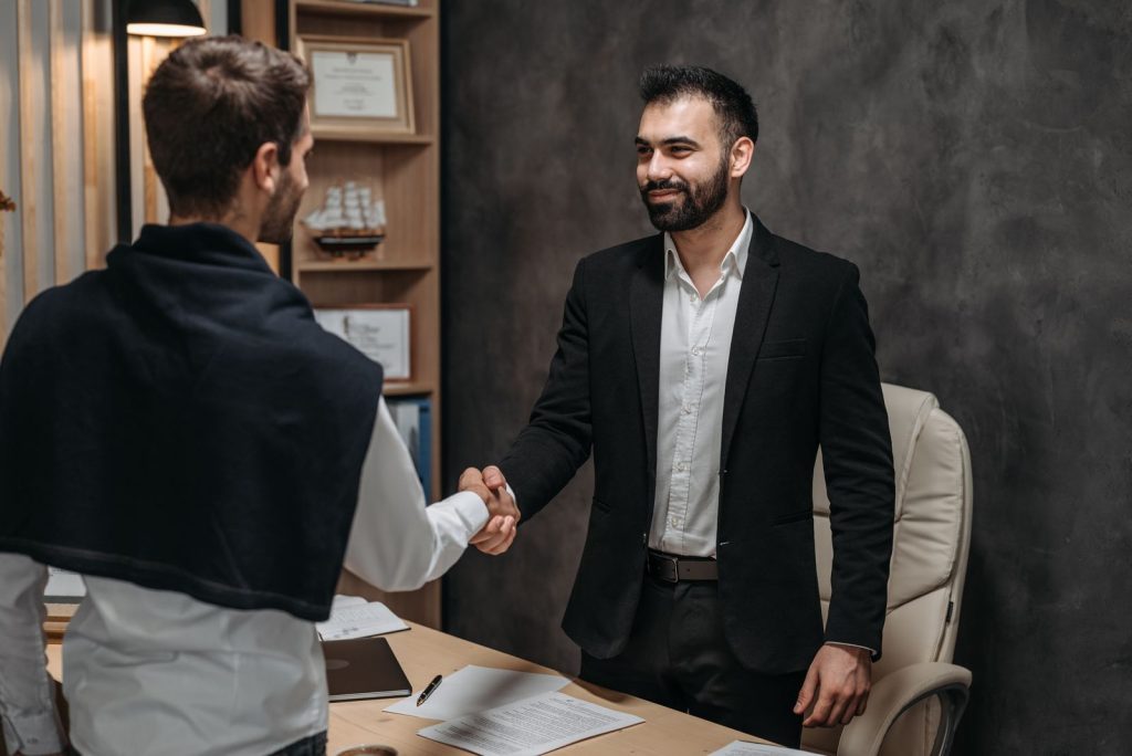 Two men shake hands in a modern office; one in a black suit smiles as they seal a deal near a wooden desk with papers and a laptop nearby.