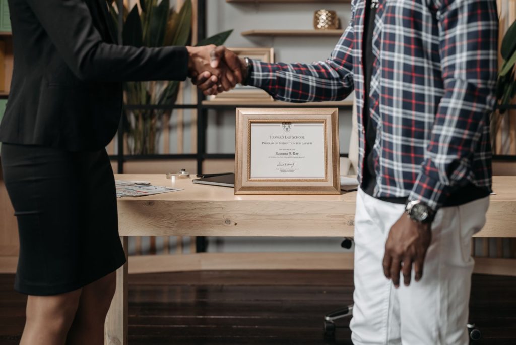 Two people shake hands across a wooden desk, with a framed certificate resting between them on the desk