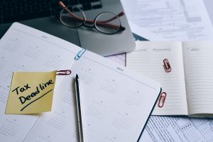 Desk scene with calendar pages, glasses resting on a laptop, and a yellow sticky note reading 'Tax time Deadlines' on top of the pages.