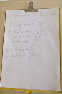 Handwritten apartment rental checklist on a clipboard listing rooms (living room, bedroom, guest room, kids room, bath, kitchen) with checked boxes