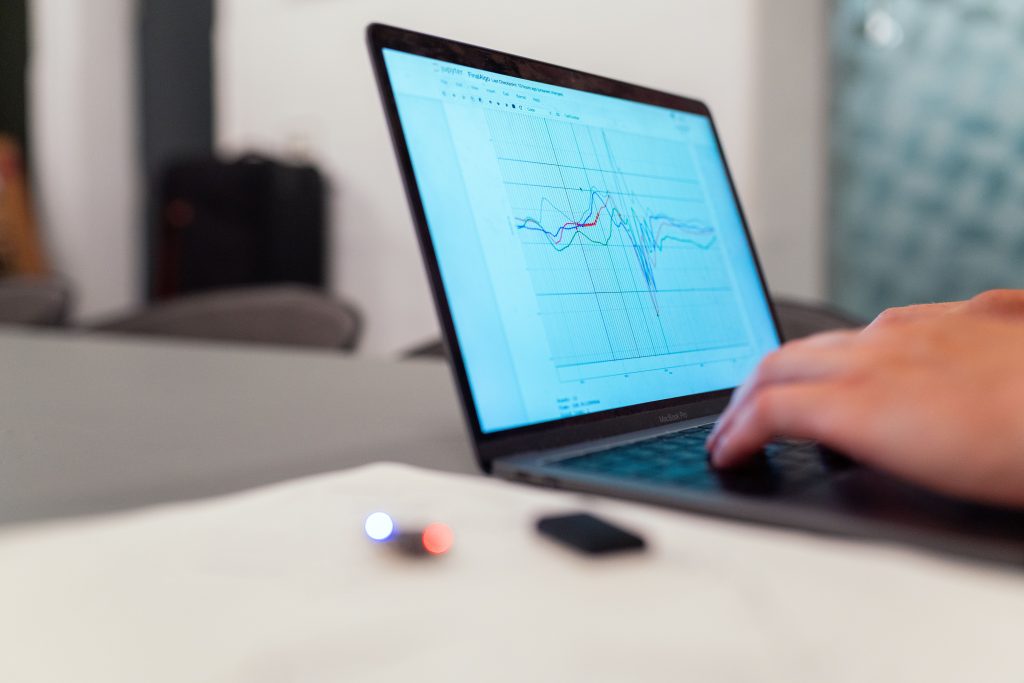 Person typing on a MacBook Pro displaying a multi-line graph with colored curves on a blue chart background, in a bright office.